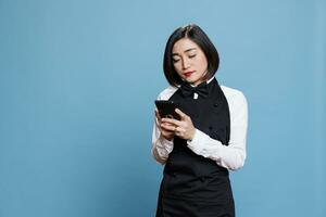 restaurante garçonete vestindo uniforme usando Smartphone para gerir cliente solicitar. jovem ásia mulher recepcionista mensagens de texto e lendo mensagem em Móvel telefone enquanto posando dentro estúdio foto