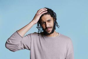 cansado árabe homem tendo dor de cabeça, tocante nuca e olhando às Câmera. Exausta jovem bonito pessoa segurando mão em cabeça e fazer face careta enquanto sofrimento a partir de enxaqueca retrato foto