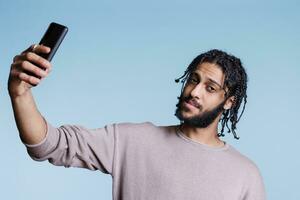 árabe homem segurando Smartphone enquanto levando selfie e olhando às Câmera. jovem pessoa vestindo casual roupas fazer foto em Móvel telefone retrato e posando em azul fundo