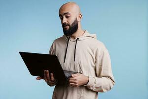 sério árabe homem segurando computador portátil e participando dentro conectados Videoconferência. jovem Careca pessoa usando portátil computador para controlo remoto conversação usando rede Video chamada Programas foto