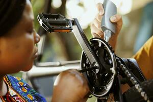 africano americano mulher examina bicicleta pedais para danos para reparar enquanto caucasiano masculino mão usa especializado lubrificante em correntes para ciclo eficiência. dois jovem ativo indivíduos trabalhando em moderno foto