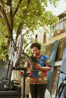 enérgico e entusiasmado Preto mulher diligentemente reparação e manutenção vários bicicleta componentes usando digital tábua dentro casa quintal. jovem desportivo fêmea surfar a Internet para manter bicicleta. foto