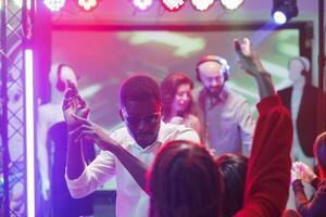 homem dançando com amigos e festa às discoteca dentro Boate. jovem africano americano clubber tendo Diversão e fazer movimentos em pista de dança às eletrônico música show dentro clube foto