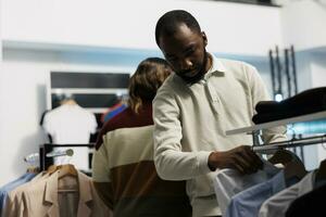 comprador escolhendo formal vestem equipamento e procurando para camisa Tamanho em prateleira dentro boutique. africano americano cliente navegando elegante vestuário em cabides enquanto compras dentro loja foto