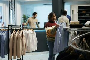 mulher segurando camisa em prateleira e olhando às Câmera enquanto escolhendo equipamento dentro roupas loja. sorridente africano americano cliente selecionando formal vestem e compras dentro boutique retrato foto