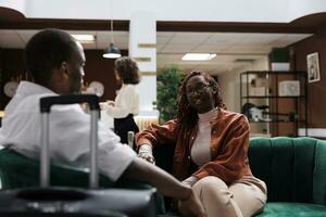 africano americano pessoas sentado dentro lobby esperando para registro às hotel recepção contador, relaxante dentro salão área. homem e mulher relaxante em sofá antes fazendo Verifica dentro procedimento. foto