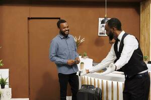 amigáveis africano americano jovem cara mensageiro dentro uniforme ajudando hotel convidado com bagagem sobre chegada, Mensageiro de hotel sorridente e acolhedor viajante dentro lobby ajudando dentro trazendo mala de viagem para dentro quarto foto