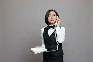 jovem confiante ásia garçonete dentro uniforme falando em telefone fixo telefone e olhando ausente. sério mulher recepcionista segurando retro Telefone e Falando com restaurante funcionários foto