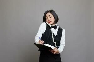 garçonete com prancheta falando em telefone fixo telefone, gerenciando restaurante operações e cliente serviço. ásia recepcionista dentro uniforme levando notas e escrevendo lista de controle enquanto conversando em Telefone foto