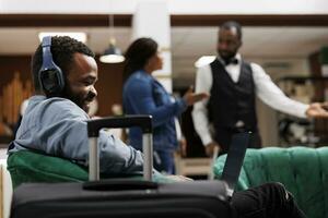 feliz africano americano homem viajante vestindo fones de ouvido olhando às computador portátil tela. hotel convidado falando para família ou amigos em vídeo ligar enquanto viajando, sentado dentro lobby usando computador foto