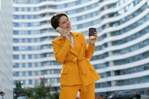 sorridente lindo mulher com curto corte de cabelo dentro amarelo Jaqueta e calça usando celular telefone , segurando copo do café . em pé sobre moderno cidade. foto