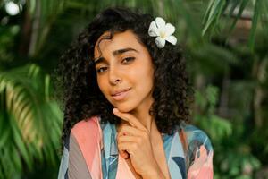 fechar acima retrato do gracioso mulher com plumeria flor dentro cabelos. vestindo boho tropical roupa. Palma árvores em fundo. foto