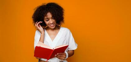 pensativo africano aluna fêmea com livro cutucando interior sobre laranja muro. foto