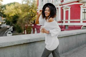 brincalhão mulher envoltórios ela mesma sobre dela ombro e olhando dentro Câmera. vestindo. à moda hipster roupa. afro Penteado . perfeito Primavera lazer Tempo dentro a tarde cidade. foto