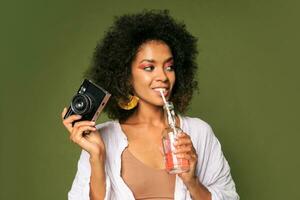 bonita africano mulher com elegante Penteado posando dentro estúdio, bebendo limonada a partir de canudo. verão estilo. verde fundo. brilhante faço acima. foto