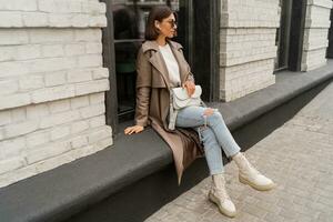 elegante elegante mulher com curto morena cabelos vestindo na moda oculos de sol, bege cor casaco, branco texturizado couro ombro bolsa, caminhando dentro rua do europeu cidade. foto