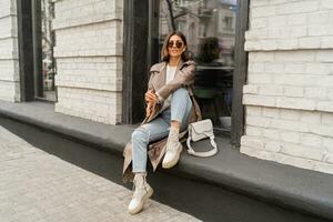 elegante elegante mulher com curto morena cabelos vestindo na moda oculos de sol, bege cor casaco, branco texturizado couro ombro bolsa, caminhando dentro rua do europeu cidade. foto