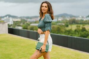 lindo morena mulher posando em cobertura topo dentro luxo hotel com visualizar. elegante acessórios, couro bolsa, à moda colheita blusa com sopro manga. foto