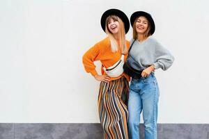 melhor amigos abraçando , casal do à moda meninas posando em branco fundo. outono temporada. vestindo à moda laranja tricotado suéter, Preto chapéu , vagabundo bolsa. amigos gastos ótimo Tempo junto. foto