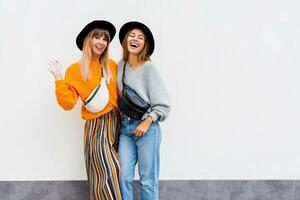 melhor amigos abraçando , casal do à moda meninas posando em branco fundo. outono temporada. vestindo à moda laranja tricotado suéter, Preto chapéu , vagabundo bolsa. amigos gastos ótimo Tempo junto. foto