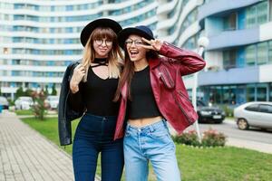 dois despreocupado sorridente mulheres posando sobre moderno cidade fundo. vestindo lã chapéu , couro Jaqueta e jeans. amigos abraçando. feliz emoções . foto
