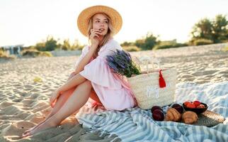 gracioso jovem mulher com loiro ondulado cabelos dentro elegante Rosa vestir desfrutando piquenique em a de praia. foto