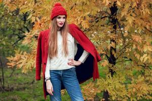 outono moda retrato do Loiras mulher dentro vermelho à moda casaco e tricotado chapéu caminhando dentro parque. foto