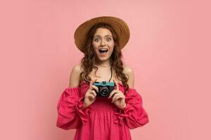 bonita vermelho cabeça fêmea turista com Câmera. estúdio tiro do espantado mulher segurando azul retro Câmera e posando sobre Rosa fundo. foto