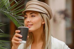 fechar acima outono retrato do romântico loiro mulher desfrutando quente café ar livre. vestindo bege na moda boina. foto