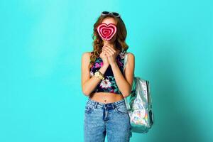 estúdio positivo retrato do jovem sexy engraçado moda louco mulher posando em azul parede fundo dentro verão estilo equipamento com Rosa pirulito vestindo impressão topo , néon mochila e fofa óculos. foto