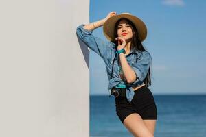 lindo morena mulher com perfeito figura dentro sexy calção e Palha chapéu posando em a de praia. azul céu e mar em fundo. foto