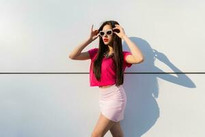 elegante morena mulher dentro verão roupas e oculos de sol posando sobre branco urbano fundo. foto