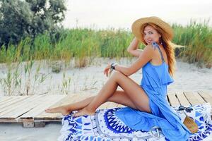 bonita fino mulher com grandes vermelho cabelos dentro Palha chapéu gastos surpreendente feriado Tempo em a de praia. vestindo azul vestir. sentado em à moda cobrir. foto