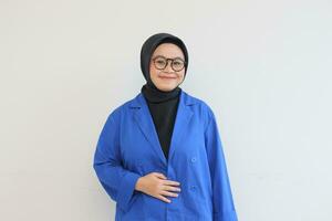 lindo jovem ásia muçulmano mulher dentro copos, hijab e vestindo azul blazer sorridente confiante e feliz com mão em barriga foto