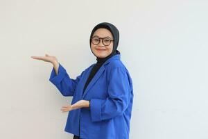 lindo jovem ásia muçulmano mulher, vestindo óculos e azul blazer mostrando Palmeiras enquanto sorridente foto