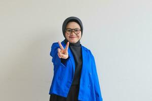 lindo jovem ásia muçulmano mulher, vestindo óculos e azul blazer gesticulando Paz símbolo foto