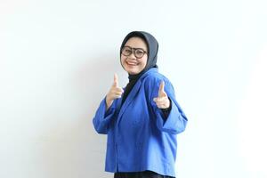lindo jovem ásia muçulmano mulher dentro copos, hijab e vestindo azul blazer sorridente confiante e animado com ambos mãos apontando frente foto