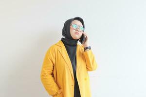 retrato do lindo ásia mulher dentro hijab, óculos e vestindo amarelo blazer fazer telefone ligar enquanto pensando foto