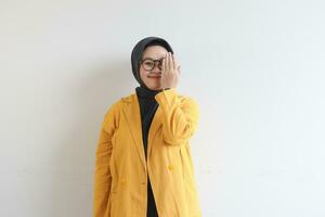 lindo jovem ásia muçulmano mulher, vestindo óculos e amarelo blazer cobertura 1 lado do face com mão enquanto sorridente foto