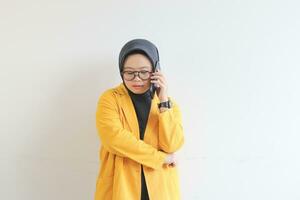 retrato do lindo ásia mulher dentro hijab, óculos e vestindo amarelo blazer fazer telefone ligar enquanto pensando foto