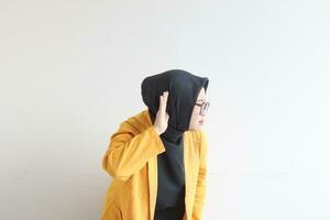 lindo jovem ásia muçulmano mulher, vestindo óculos e amarelo blazer com mão gesto ouvindo para alguma coisa ou alguém foto