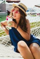 atraente jovem mulher dentro azul vestir e Palha chapéu vestindo Rosa oculos de sol bebendo coquetéis foto