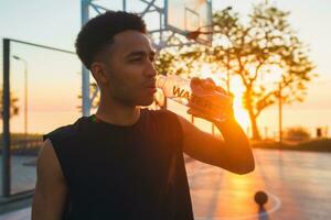 Preto homem fazendo Esportes dentro manhã, bebendo água em basquetebol quadra em nascer do sol foto