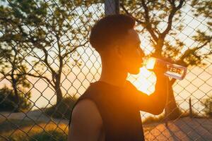 Preto homem fazendo Esportes dentro manhã, bebendo água em basquetebol quadra em nascer do sol foto