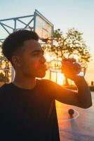 Preto homem fazendo Esportes dentro manhã, bebendo água em basquetebol quadra em nascer do sol foto
