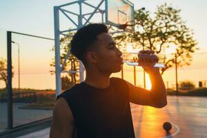 Preto homem fazendo Esportes dentro manhã, bebendo água em basquetebol quadra em nascer do sol foto