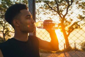 Preto homem fazendo Esportes dentro manhã, bebendo água em basquetebol quadra em nascer do sol foto