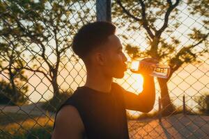 Preto homem fazendo Esportes dentro manhã, bebendo água em basquetebol quadra em nascer do sol foto