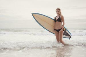 retrato do sorridente jovem mulher em pé com prancha de surfe às a de praia foto