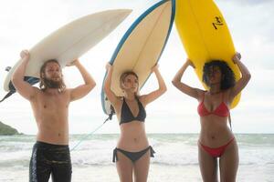traseiro Visão do dois mulheres e jovem homem segurando pranchas de surf em seus cabeças e andar para dentro a mar para surfar foto